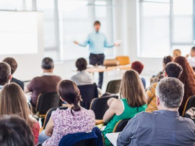 Speaker giving presentation on business conference. Business and entrepreneurship symposium. Speaker giving a talk at business meeting. Audience in conference hall. Rear view of unrecognized participant in audience.